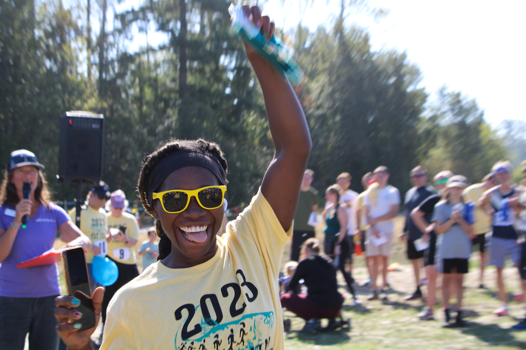 A joyful person with sunglasses and a "2023" shirt raises their arm at an outdoor event, surrounded by a crowd.