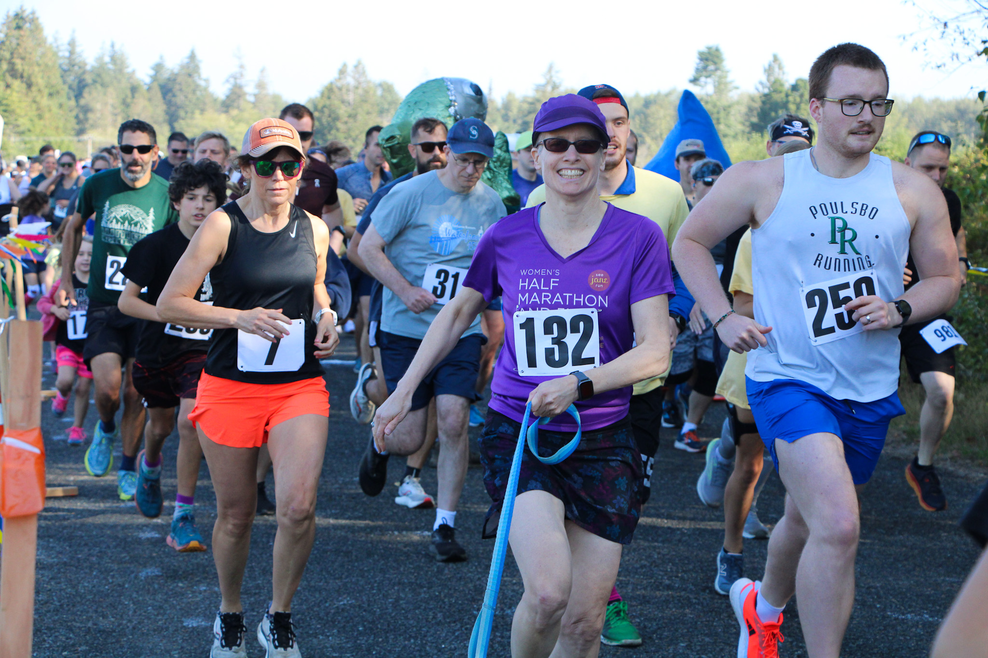 A group of runners in a race, some wearing event bibs. They're on a road with trees in the background.