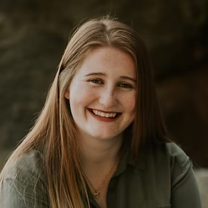 A person with long hair smiling, wearing a dark top, in front of a blurred outdoor background.
