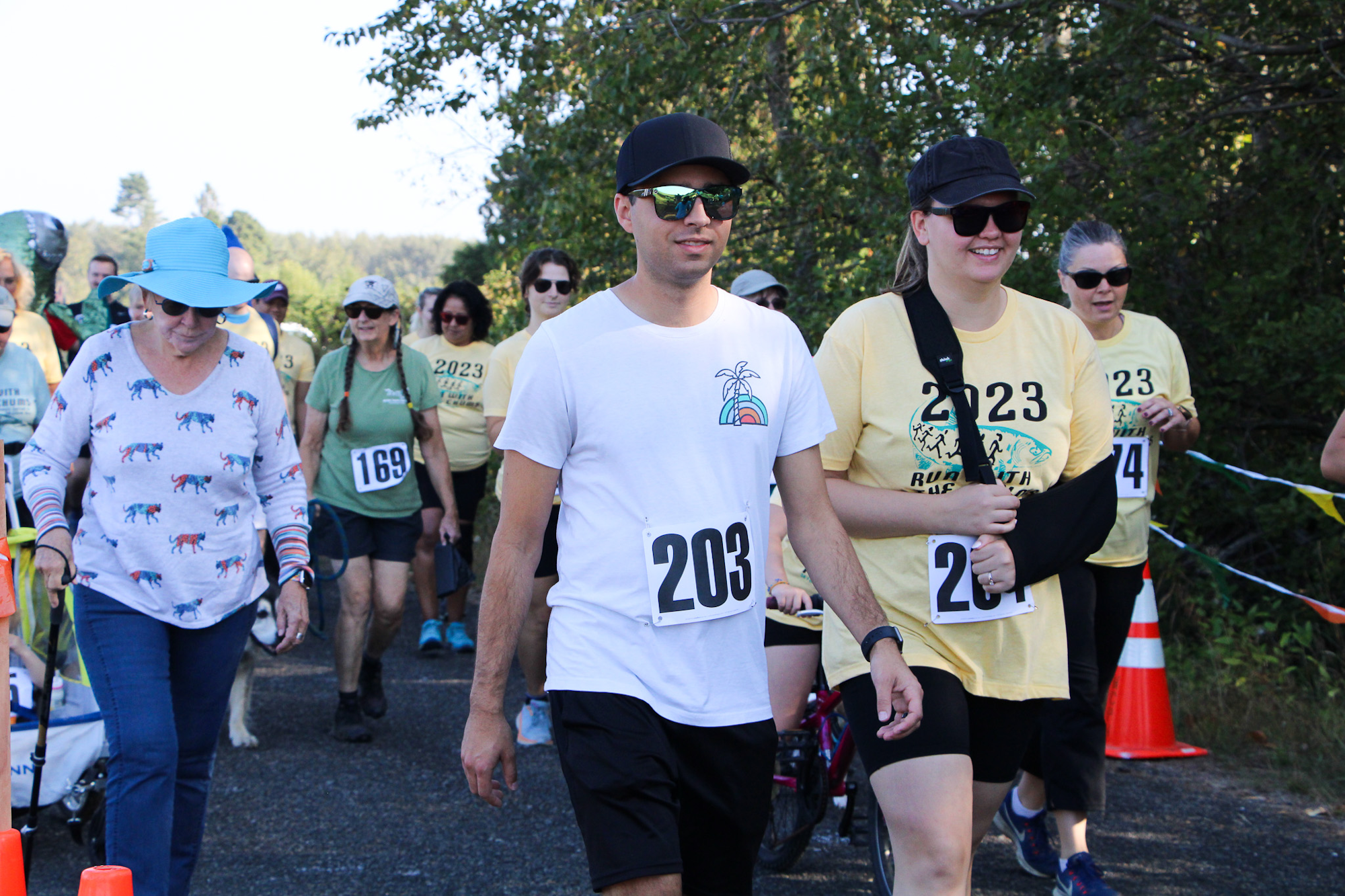 People participating in an outdoor race or walk event, wearing numbered bibs and casual clothing, with trees and a path in the background.