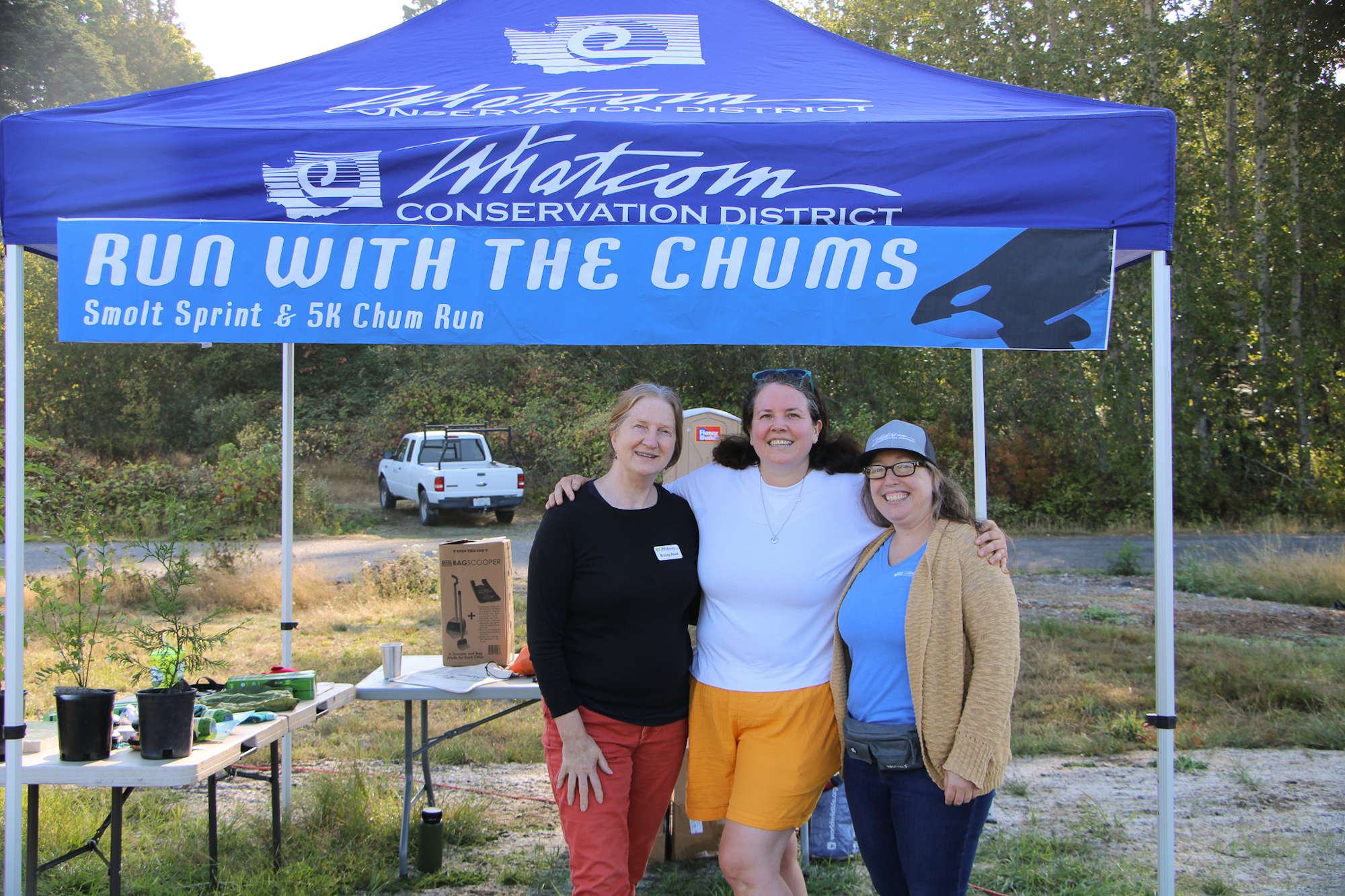 Three people under a "Run with the Chums" banner at a Whatcom Conservation District event.