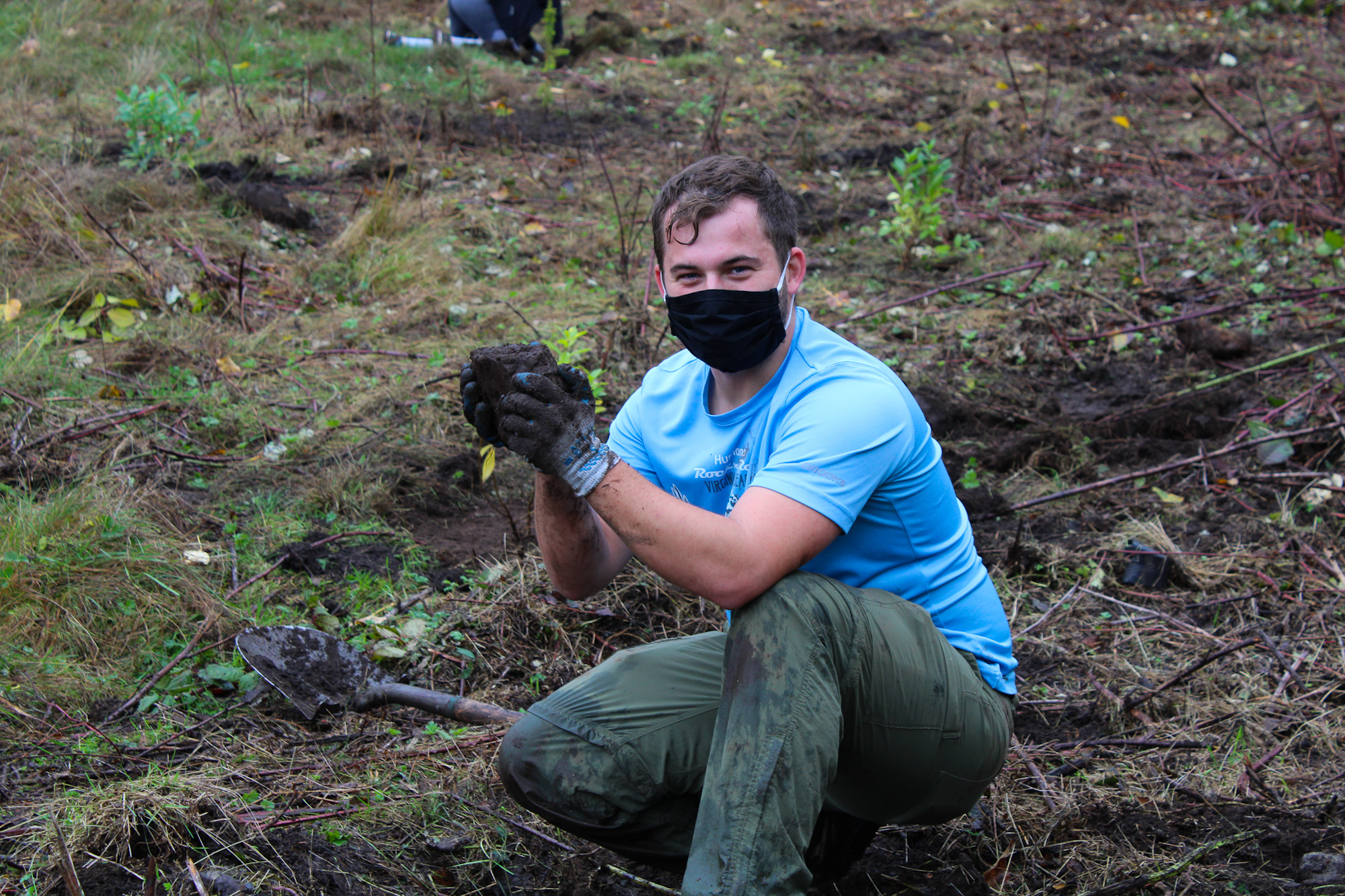 A person wearing a black mask and gloves, kneeling on grass, holding soil with a shovel nearby.