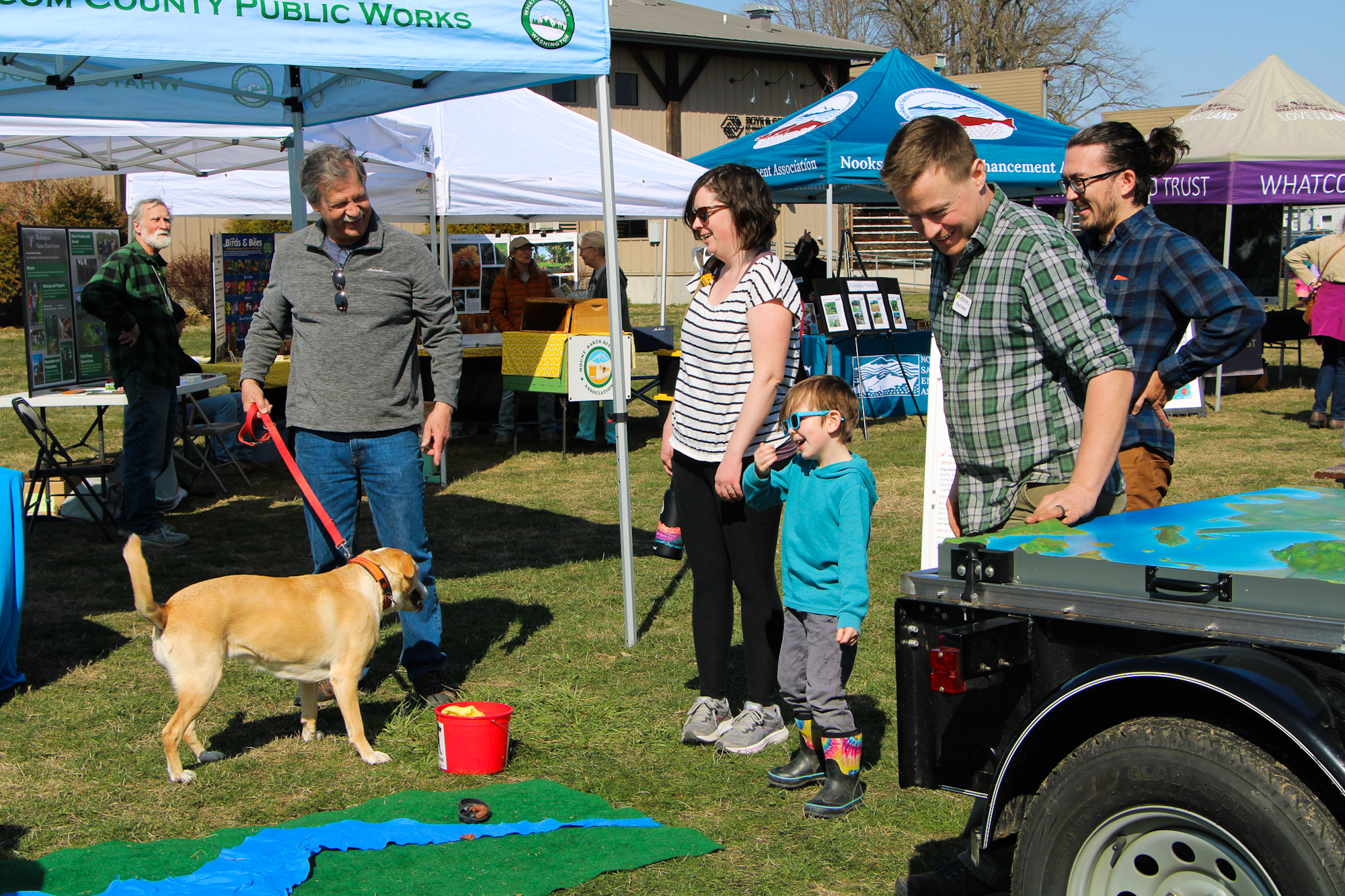 Outdoor event with tents, people, a dog on a leash, and a child in sunglasses.
