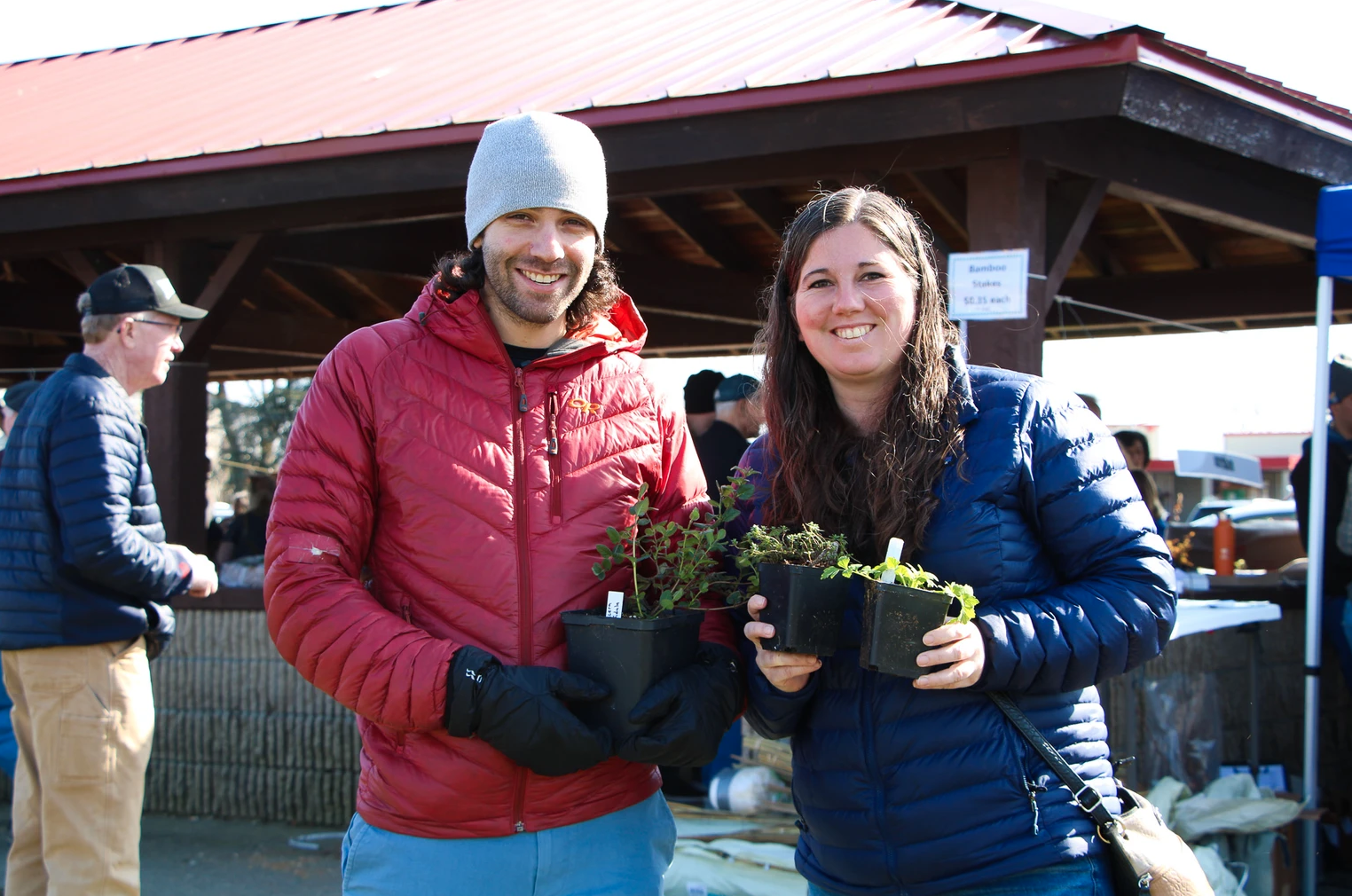 Two people smiling, holding potted plants at an outdoor market or fair.