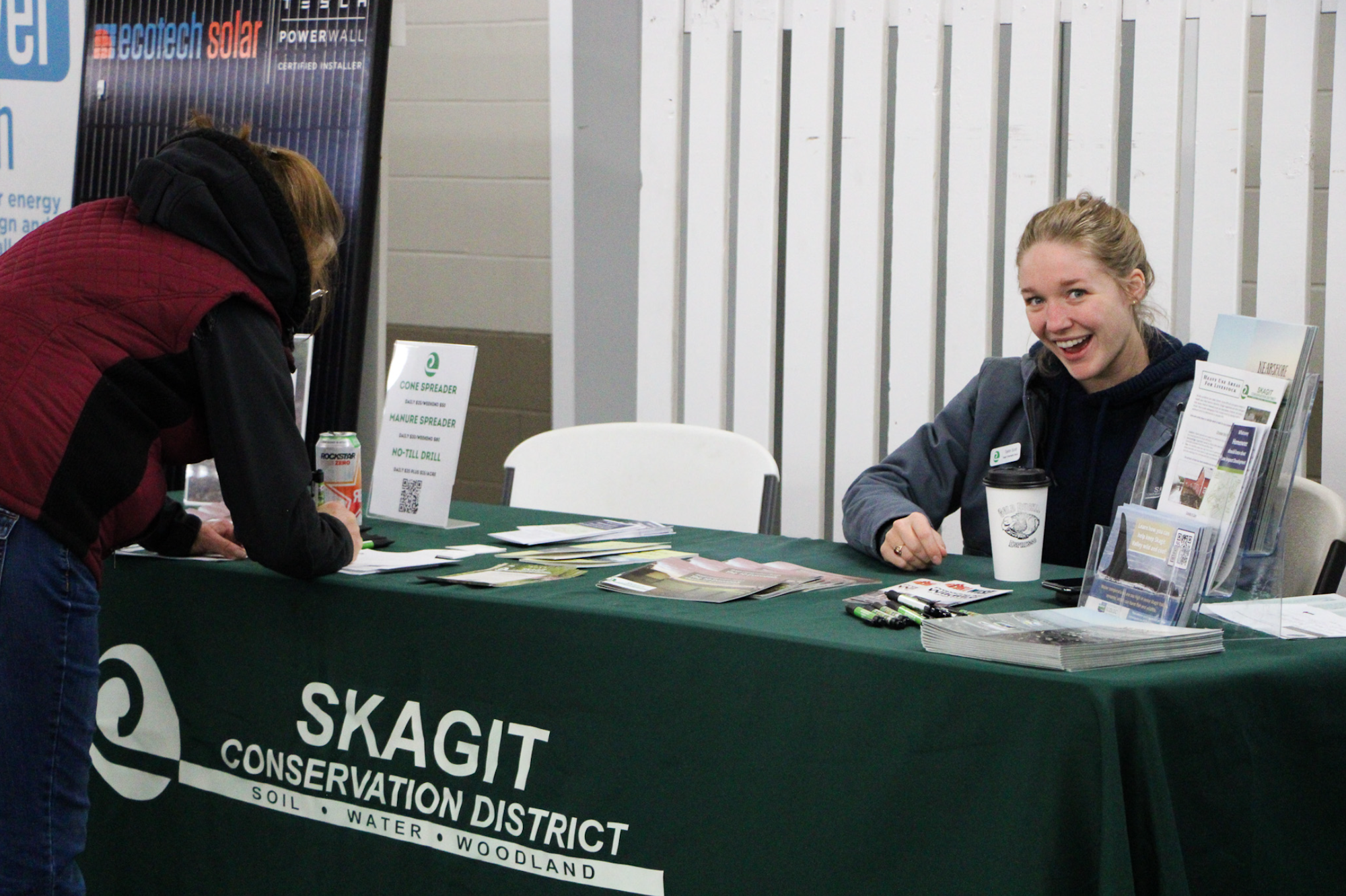 Two people at a booth for Skagit Conservation District. One is sitting and smiling; the other is writing.