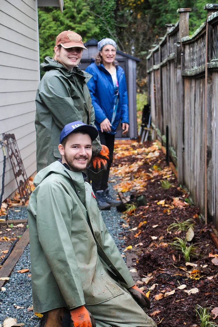 Three people in outdoor clothing, smiling while gardening beside a house and fence.