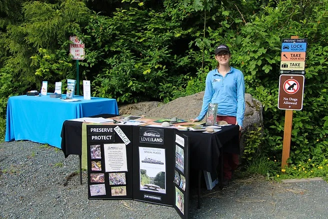 A person stands by a table with informational displays about preservation and protection, set in a lush outdoor area.