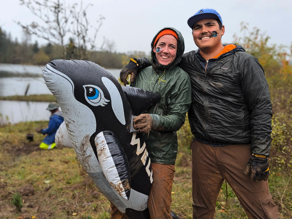 Two people outdoors, muddy, with an inflatable orca, smiling by a lake.