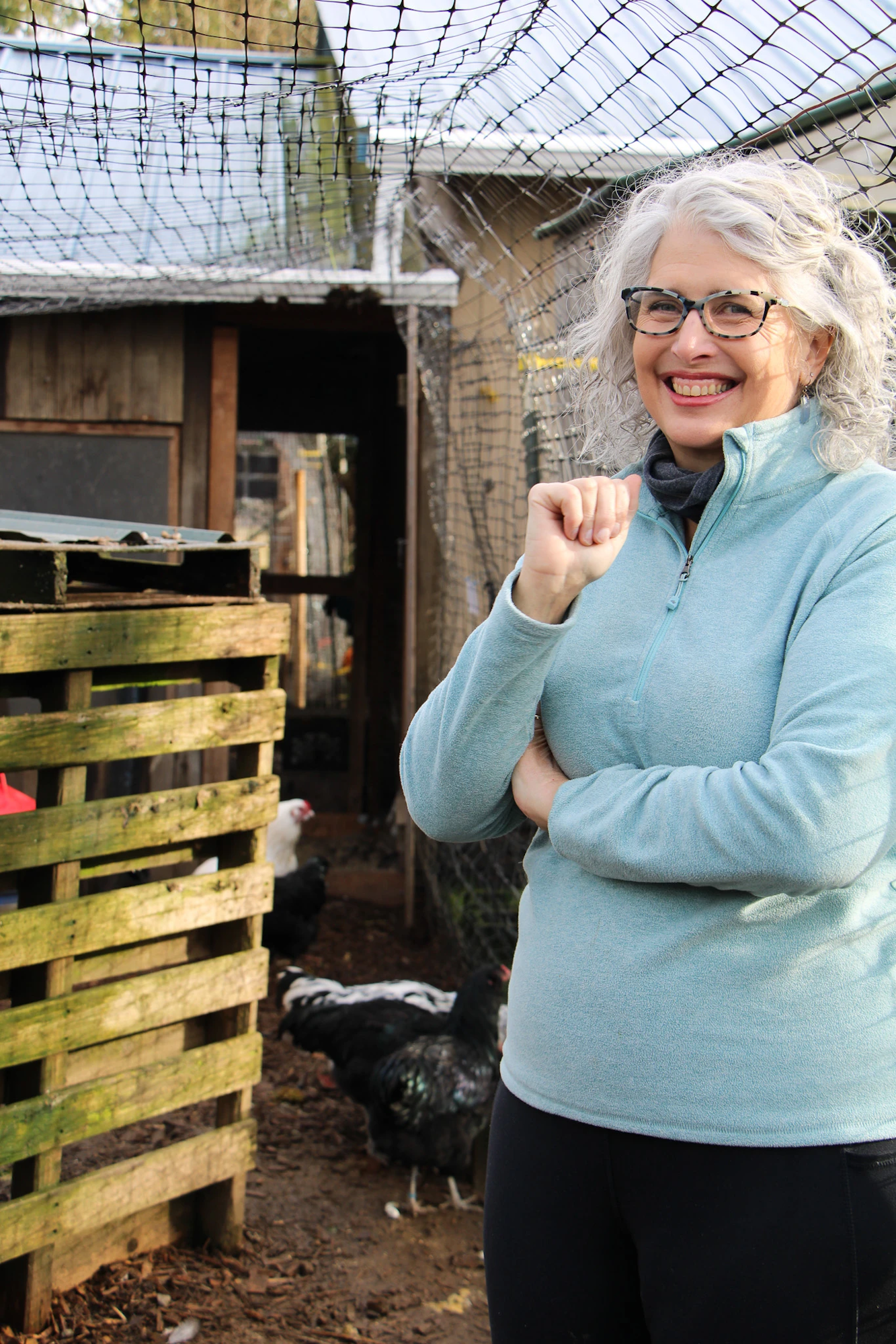 A smiling person in glasses and a blue sweater stands next to a chicken coop with chickens and a netted roof.