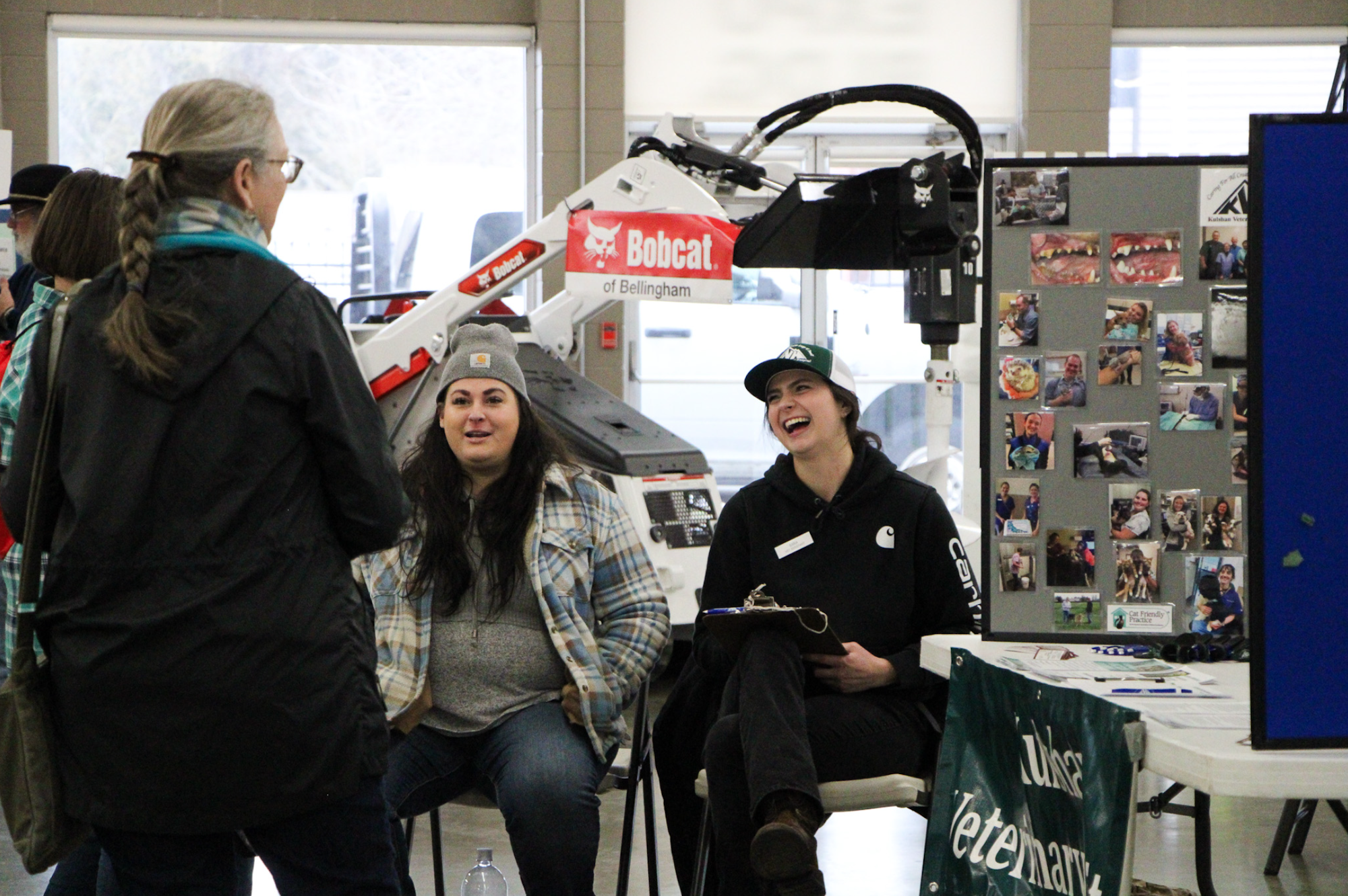 People interacting at an event with photos on a board and a Bobcat machine in the background.