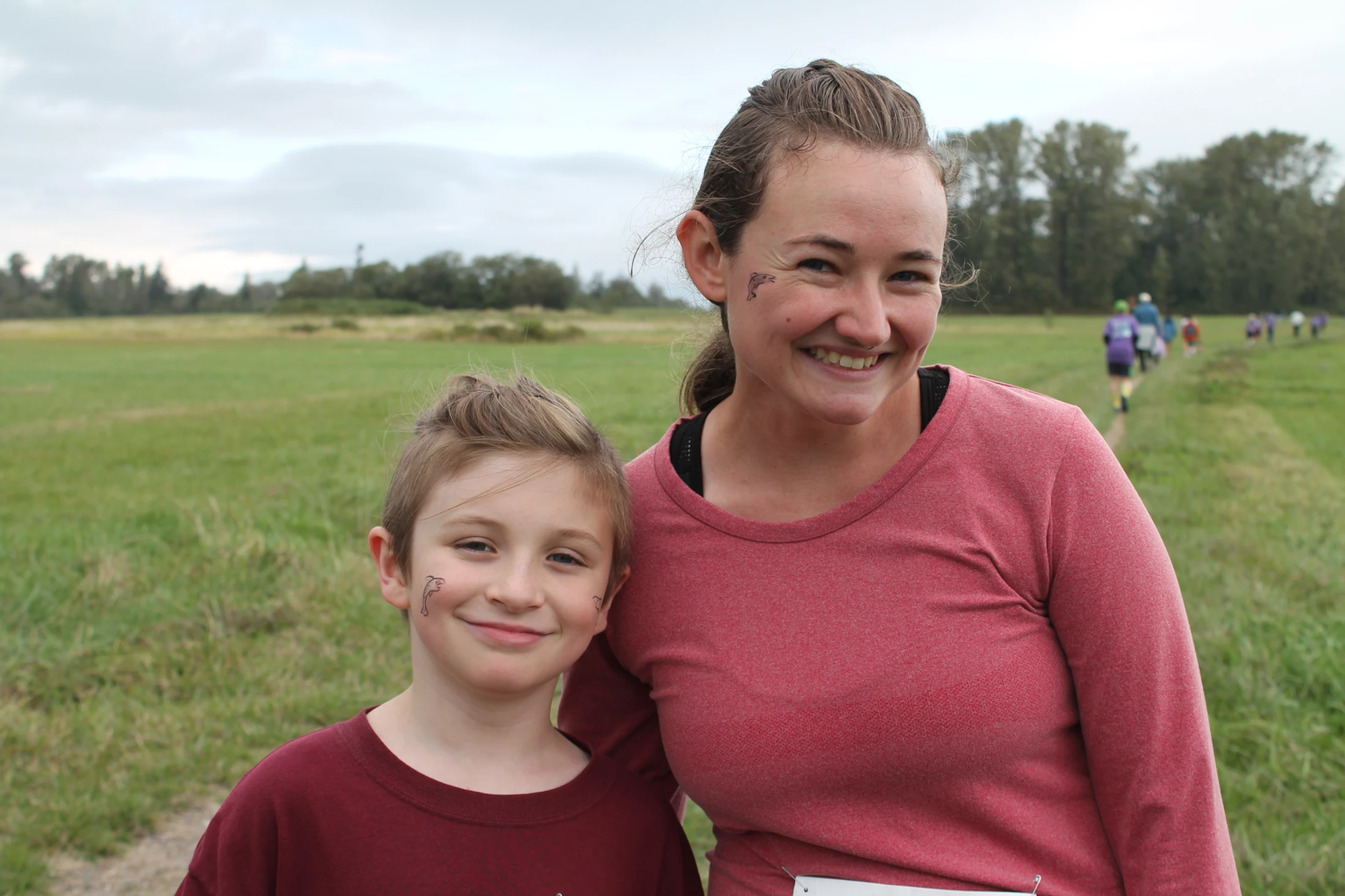 A woman and a child smiling outdoors, wearing maroon tops, with grass and trees in the background.