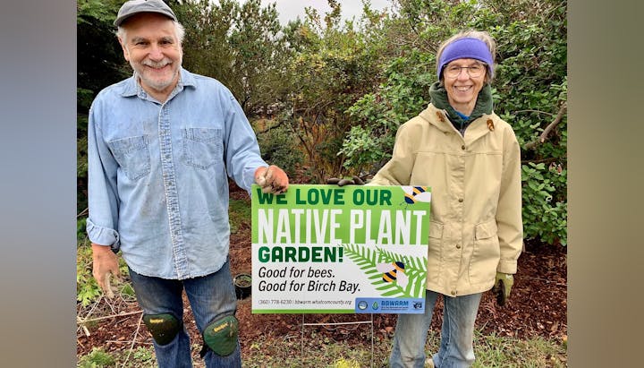 Two people holding a sign that says "WE LOVE OUR NATIVE PLANT GARDEN!" with contact info and logos.