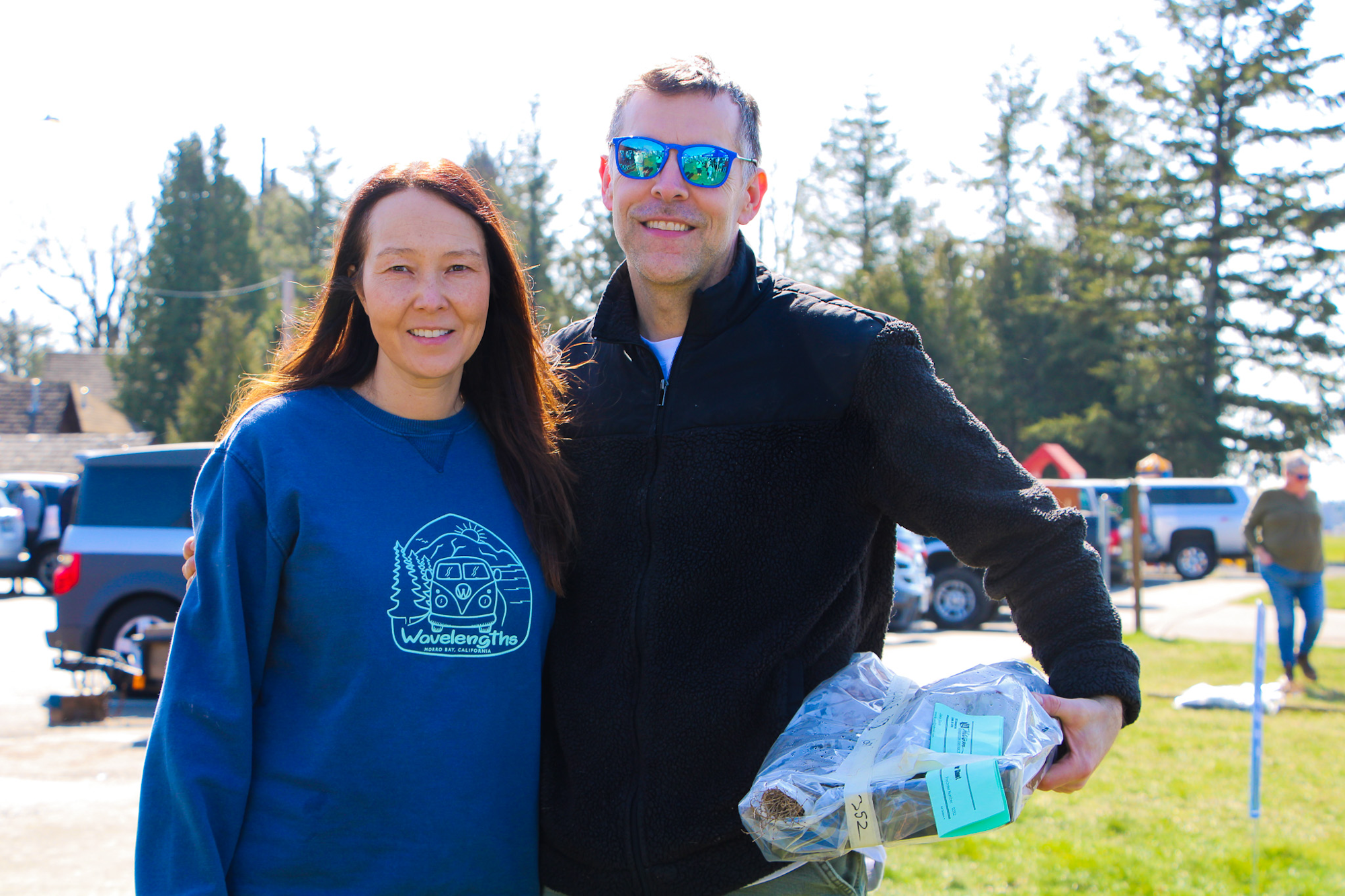 Two people posing outdoors, one holding packaged items, with trees and vehicles in the background.
