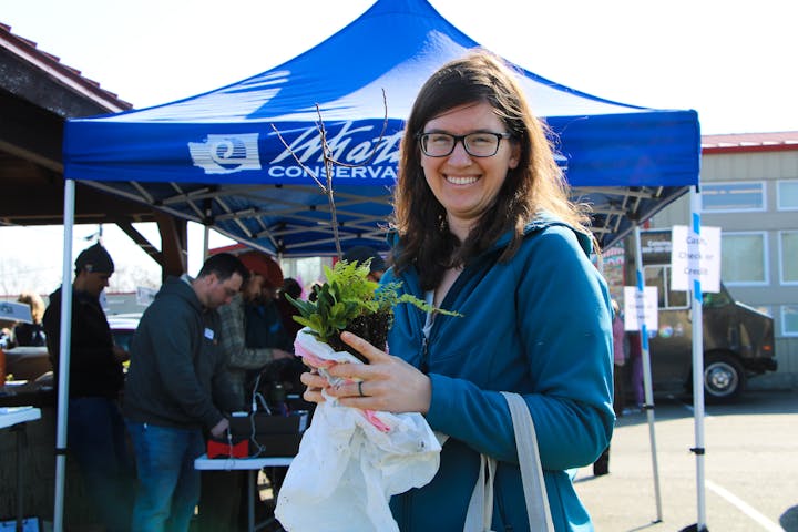 A woman smiling, holding plants in front of a blue tent at an outdoor event.