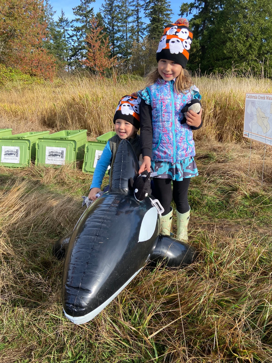 Two kids in hats with ghosts stand by an inflatable orca in a field, surrounded by green bins.