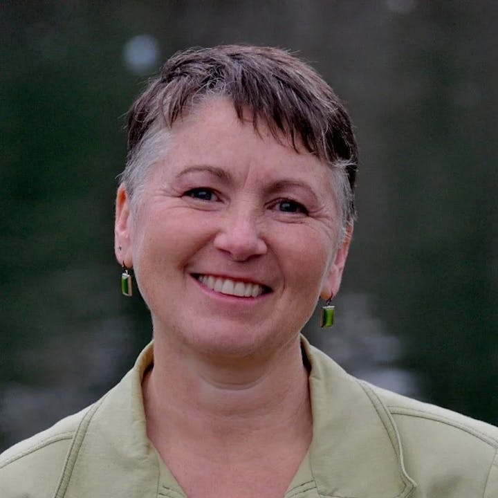 A smiling person with short hair and green earrings against a blurred dark background.