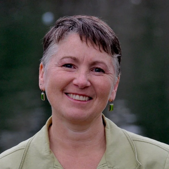 A smiling person with short hair and green earrings against a blurred dark background.