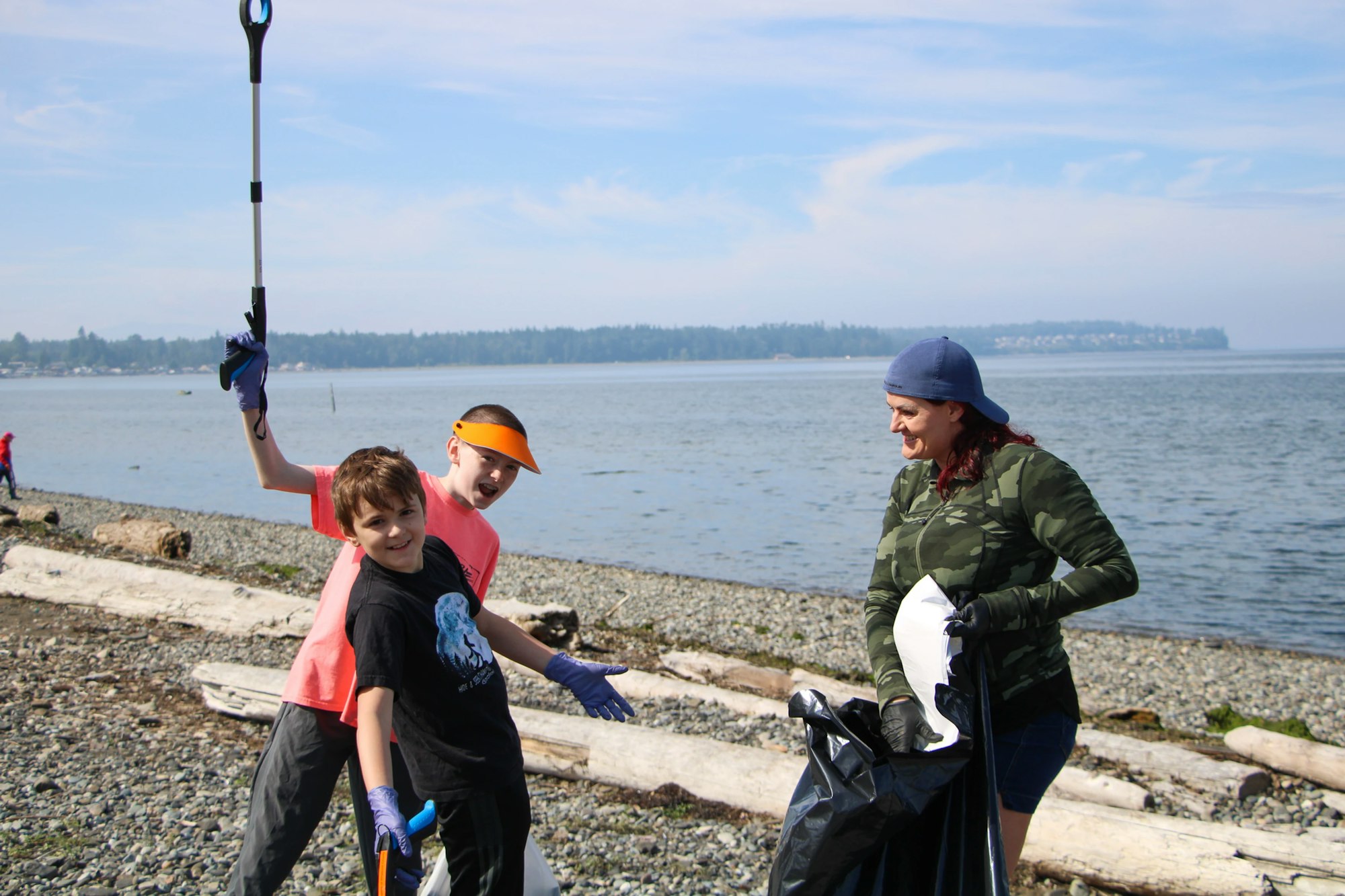 Three people on a beach picking up trash, two kids and an adult, using gloves and grabbers, with the ocean in the background.
