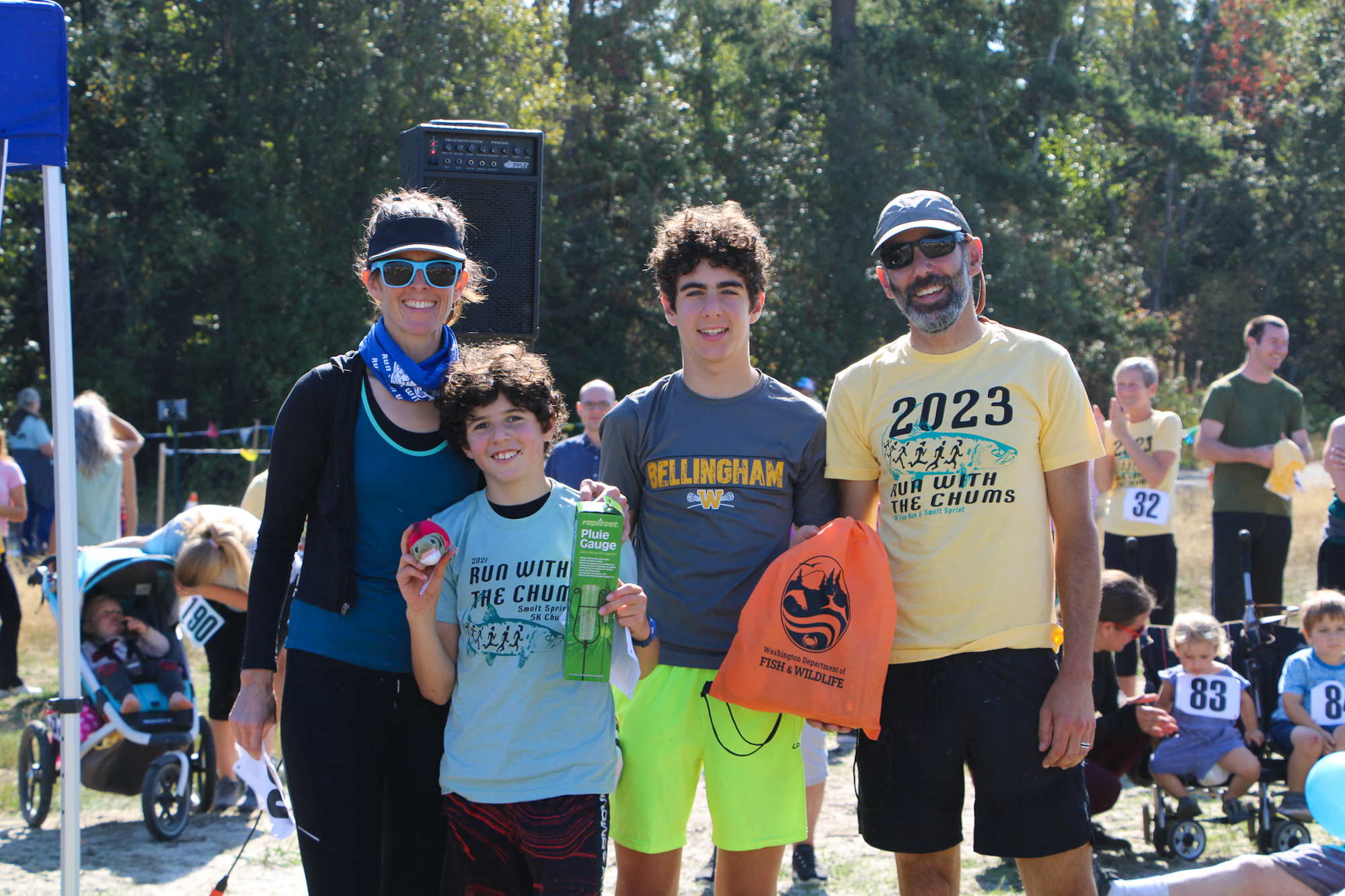 A group of four people smiling at an outdoor event, holding prizes and wearing "Run With the Chums" shirts.