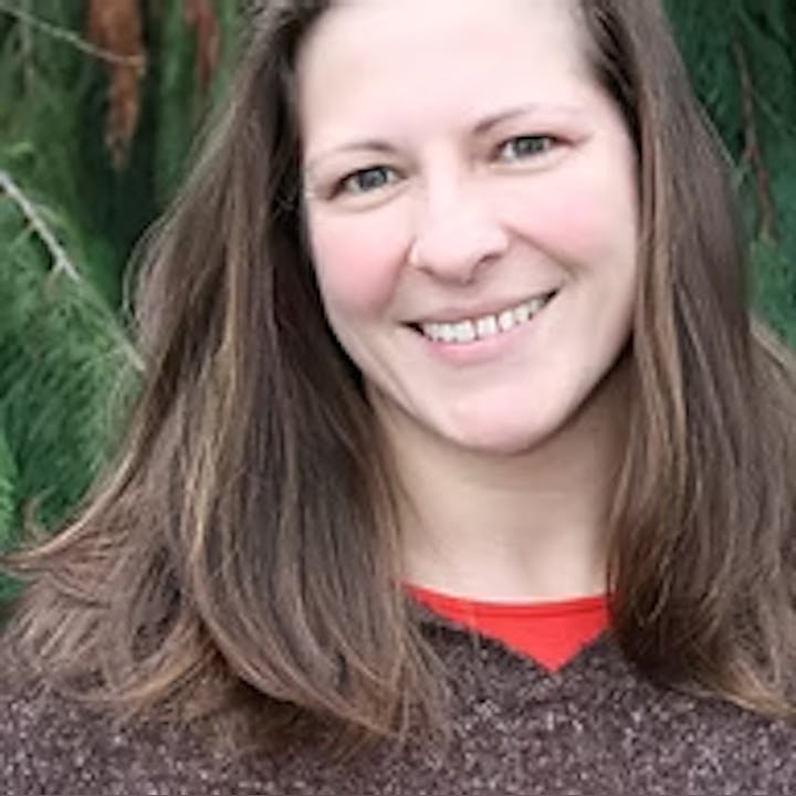 A person with long brown hair smiling outdoors in front of green foliage.
