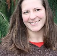A person with long brown hair smiling outdoors in front of green foliage.