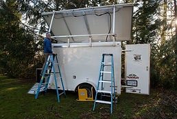 A person stands on a ladder next to a trailer-like structure with open panels and equipment, surrounded by trees.
