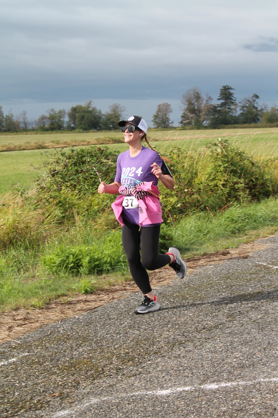 Person jogging outdoors on a path, wearing a purple shirt and black leggings, with a race number 37. Grass and trees in the background.