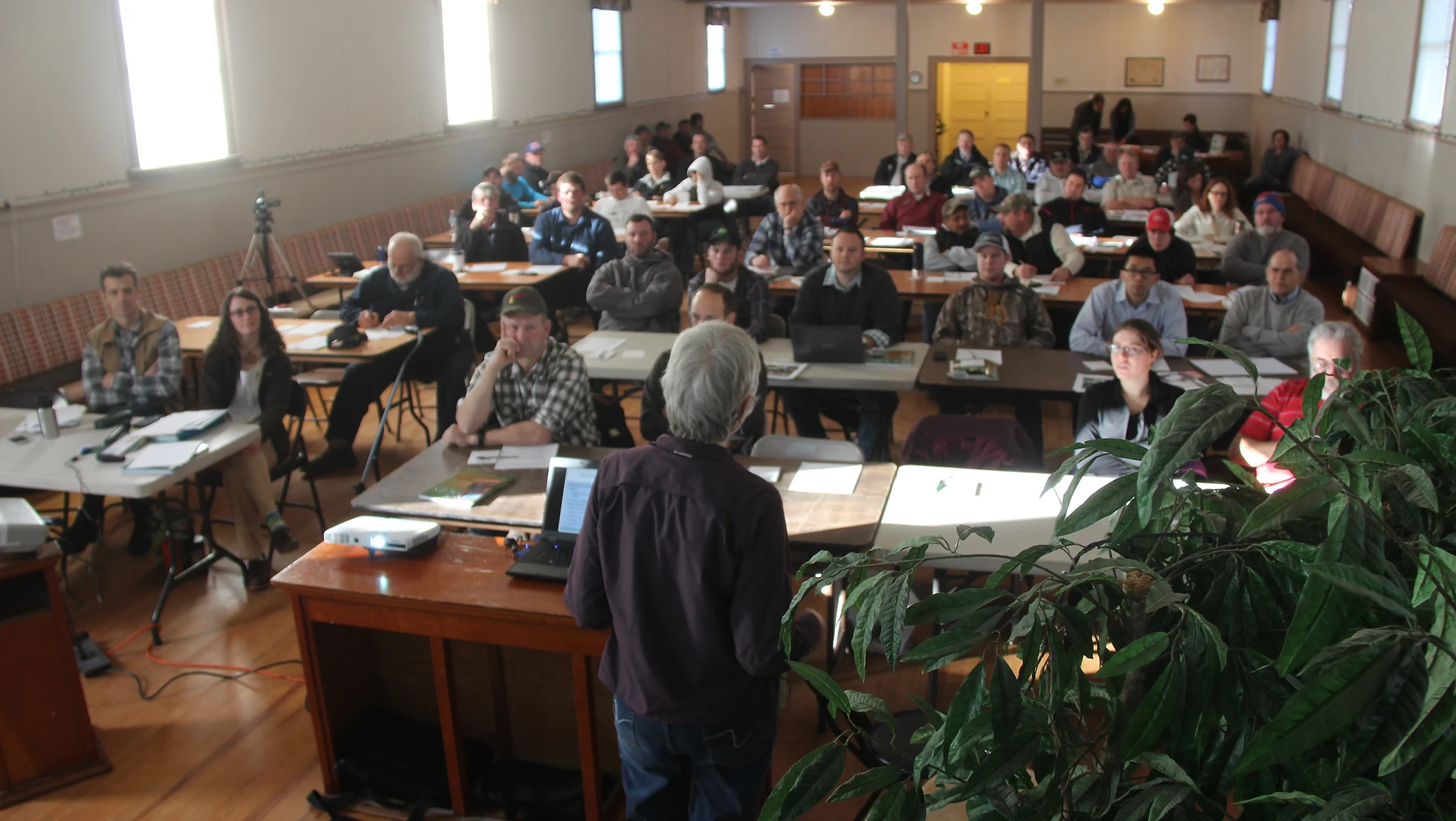 A person presents to a room filled with attentive individuals. Tables have laptops and papers, and plants are visible in the foreground.