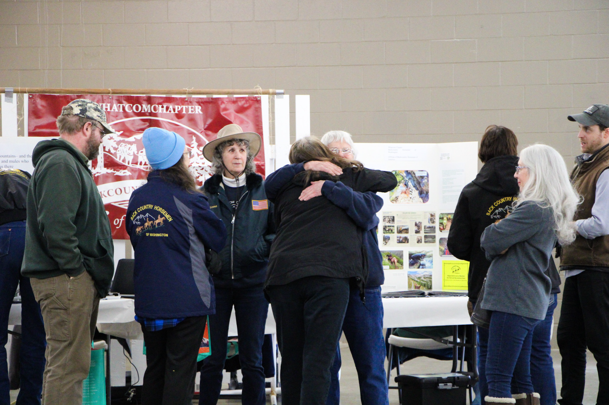 A group of people are gathered indoors, some talking and two hugging, in front of display boards and a banner.