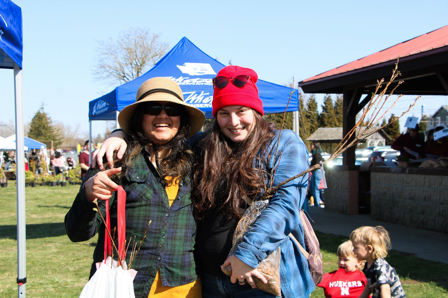 Two people smiling at an outdoor event, holding small plants, with tents and other attendees in the background.