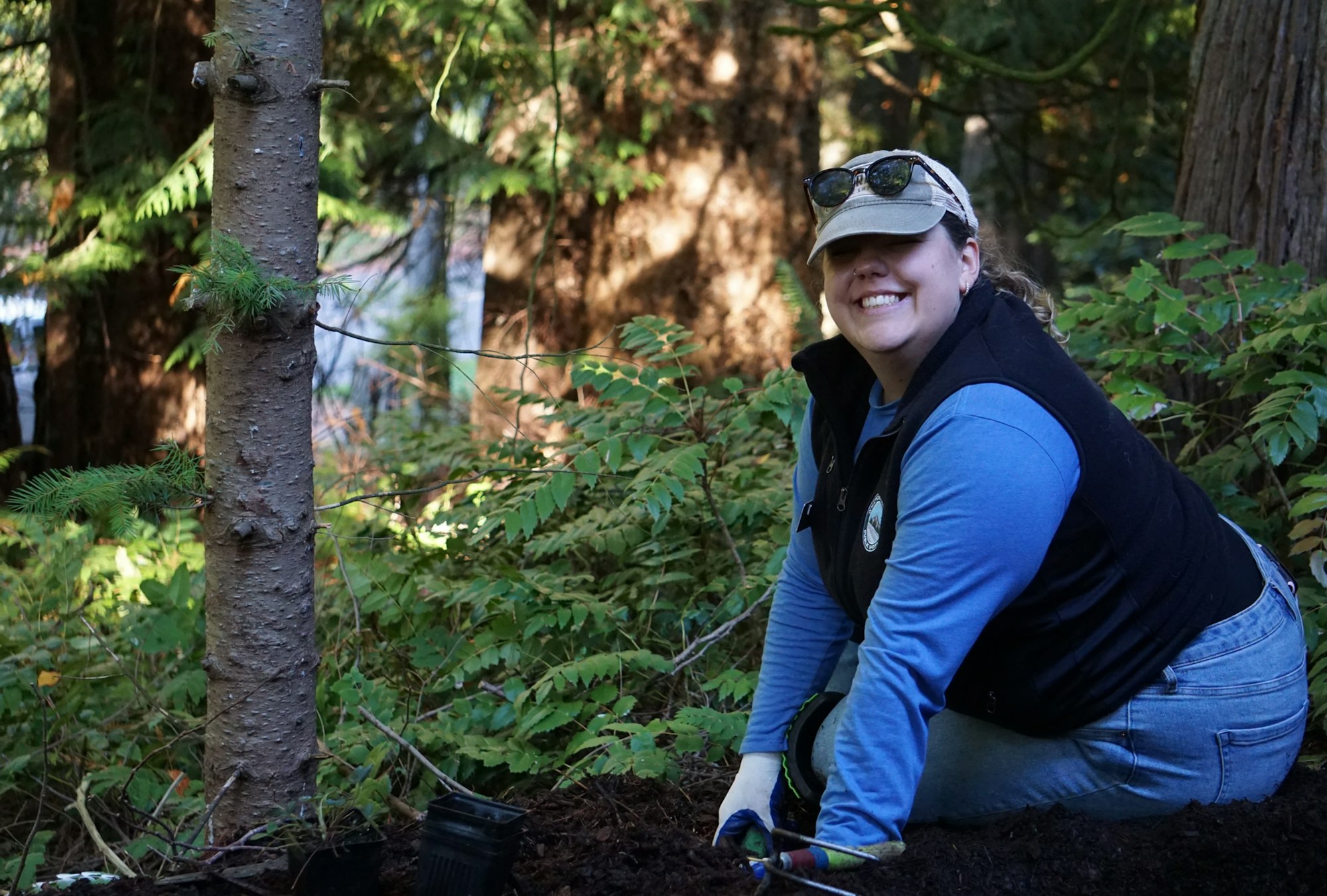 A person smiles while gardening in a forested area, wearing gloves, a cap, and sunglasses on top.