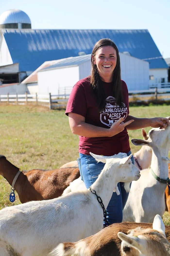 A person smiling while standing among goats in front of a barn.