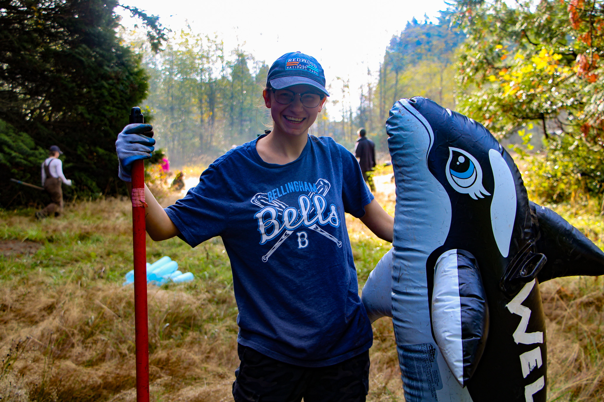 A person smiling, wearing a hat and holding a tool, is next to an inflatable orca in a grassy outdoor area.