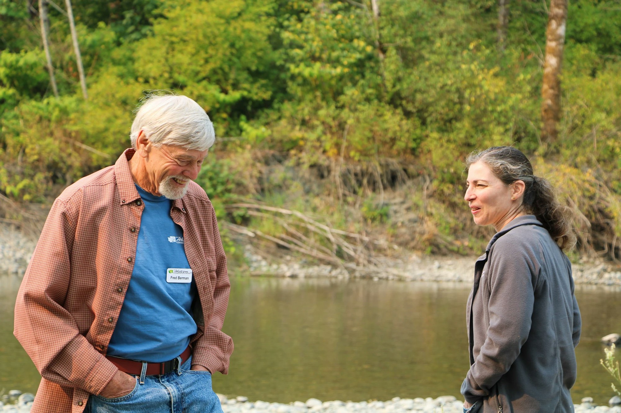Two people smiling and talking by a river, with a forested background.