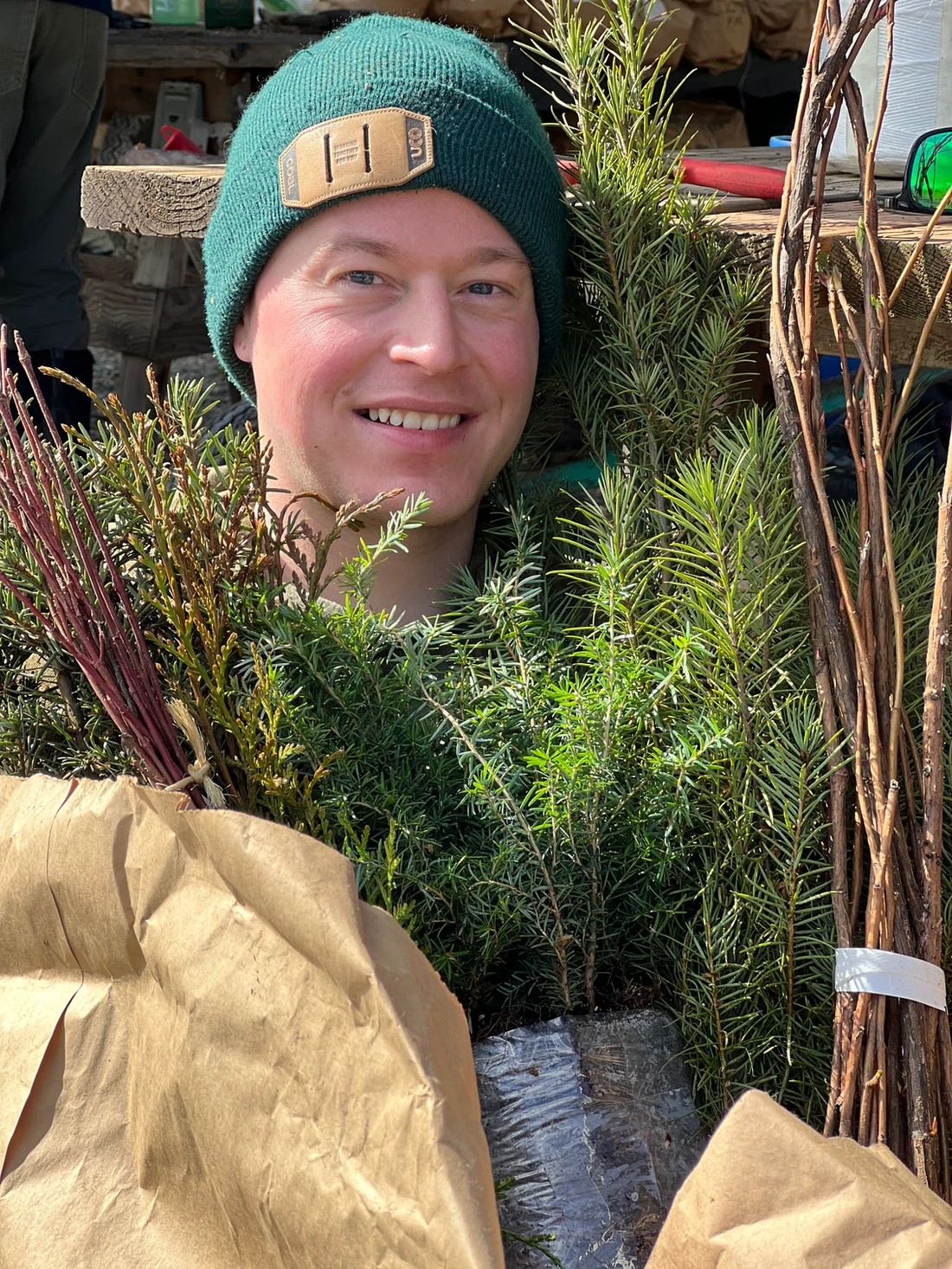 A smiling person in a green beanie surrounded by various green plants and branches, with paper wrapping visible in the foreground.