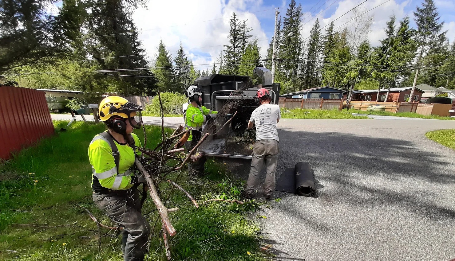 Workers in safety gear load branches into a chipper on a residential street.