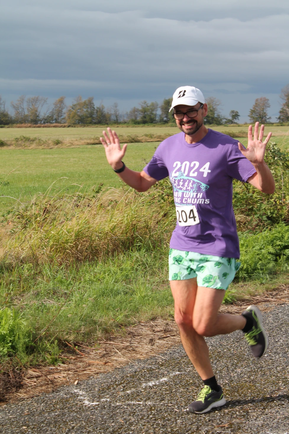 A person running in a race outdoors, wearing a purple shirt, green shorts, and a white cap, smiling with both hands raised.