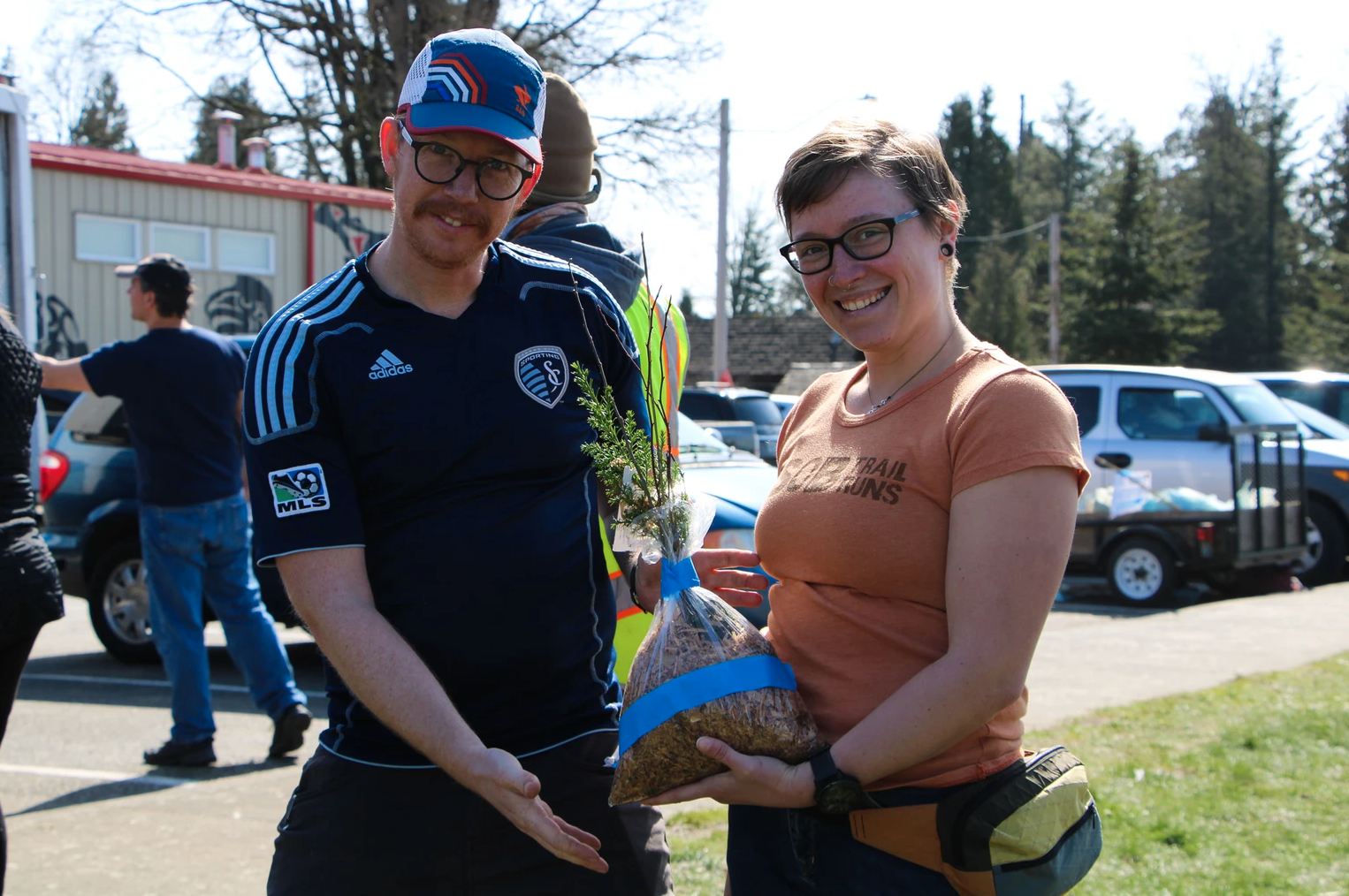 Two people smiling, holding a small tree in a bag, outdoors near parked cars.