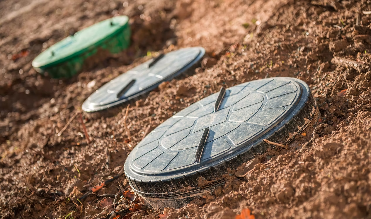 Three septic tank lids in a row, partially buried in the ground.