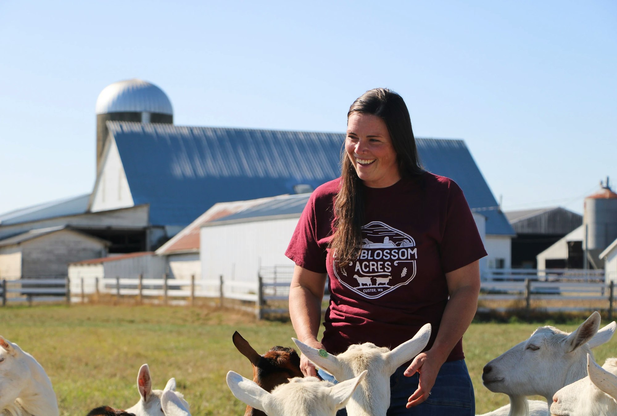 A person smiling on a farm with goats, wearing a T-shirt that says "Blossom Acres," in front of barn buildings.