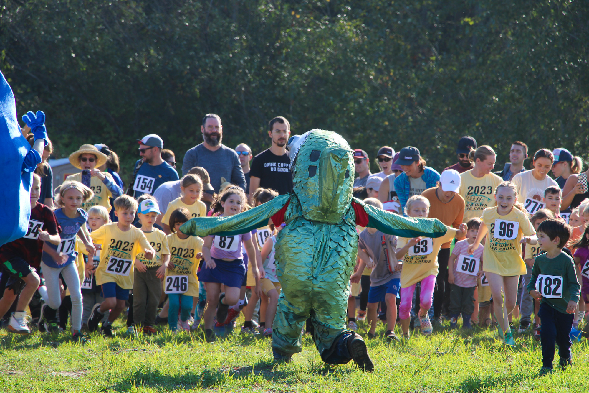 Kids and adults running in a race, led by a person in a green costume, outdoors.