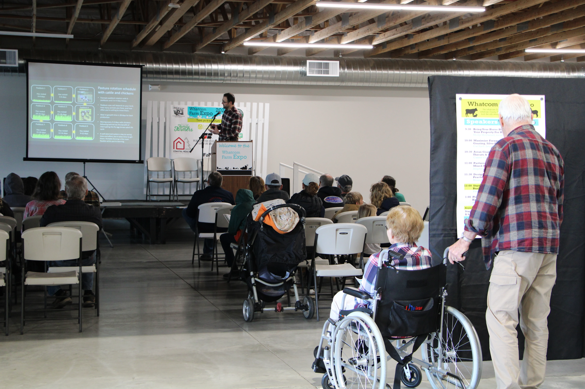 A presentation at Whatcom Farm Expo with an audience seated, including a person in a wheelchair and a schedule board displayed.