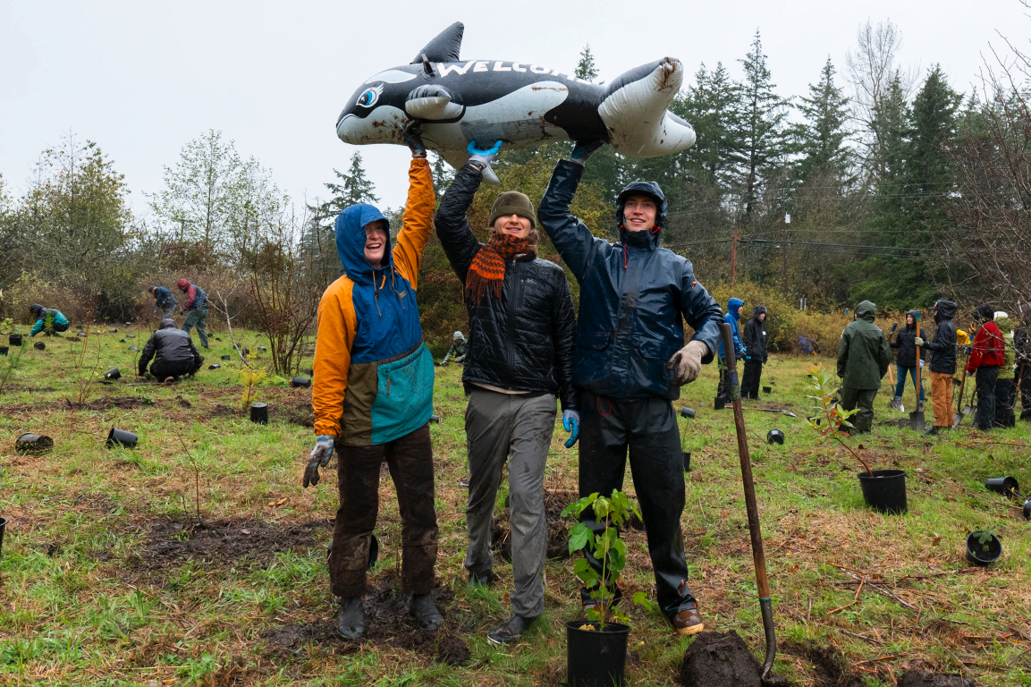 People planting trees, smiling, and holding a "Welcome" inflatable animal in a grassy area with scattered potted plants.