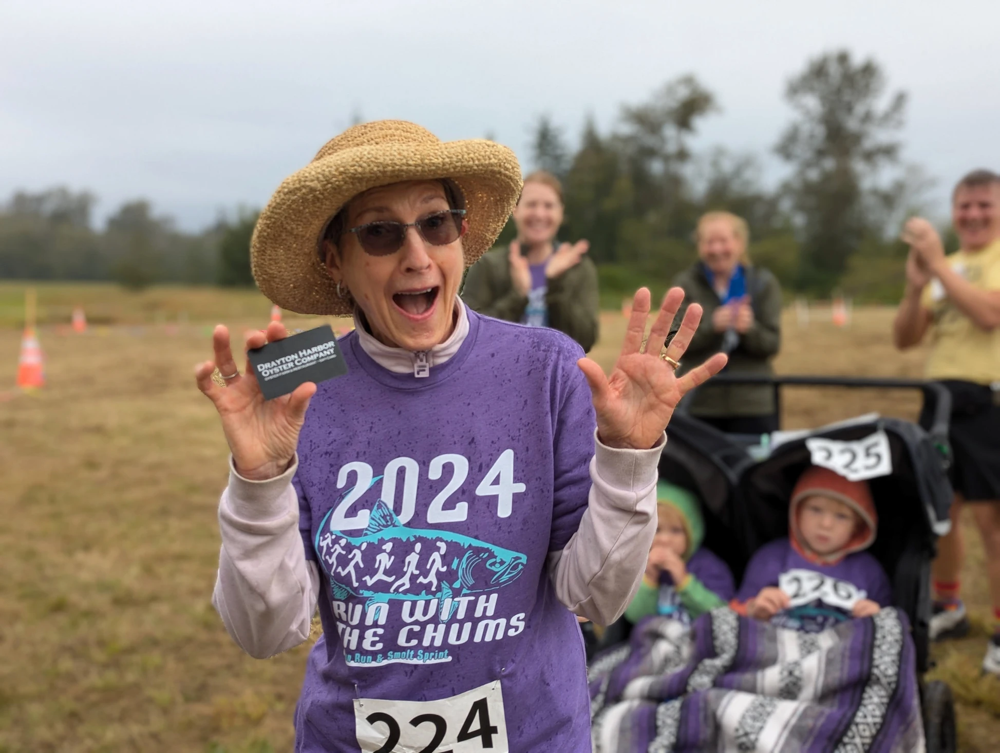 Person in a purple shirt and sun hat excitedly holds a card; others clap in the background.