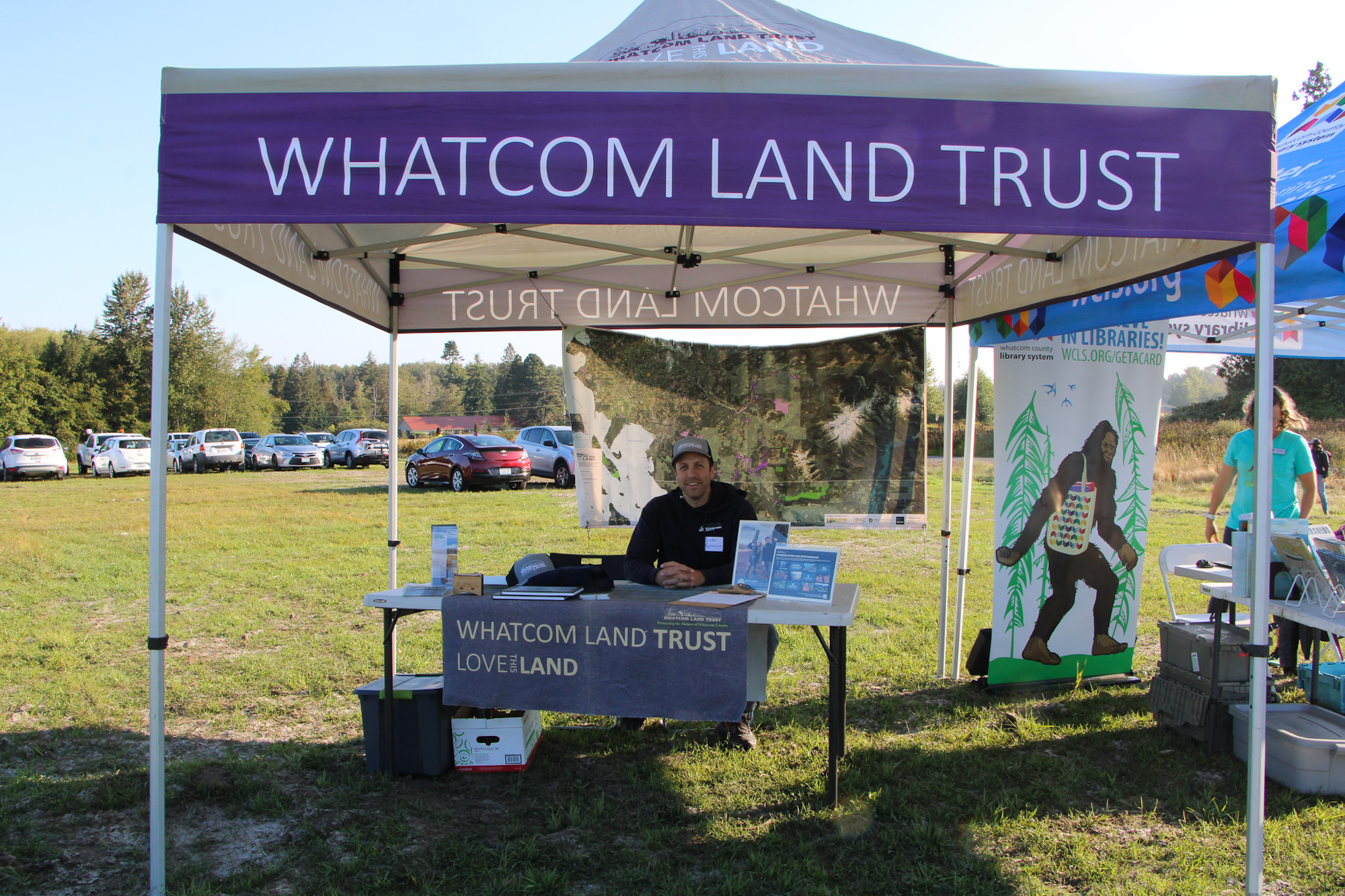 A Whatcom Land Trust booth with a man at a table, surrounded by maps and informational materials, set outdoors.