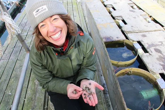 A smiling person in a beanie and jacket holding small marine creatures on a dock with circular tanks.