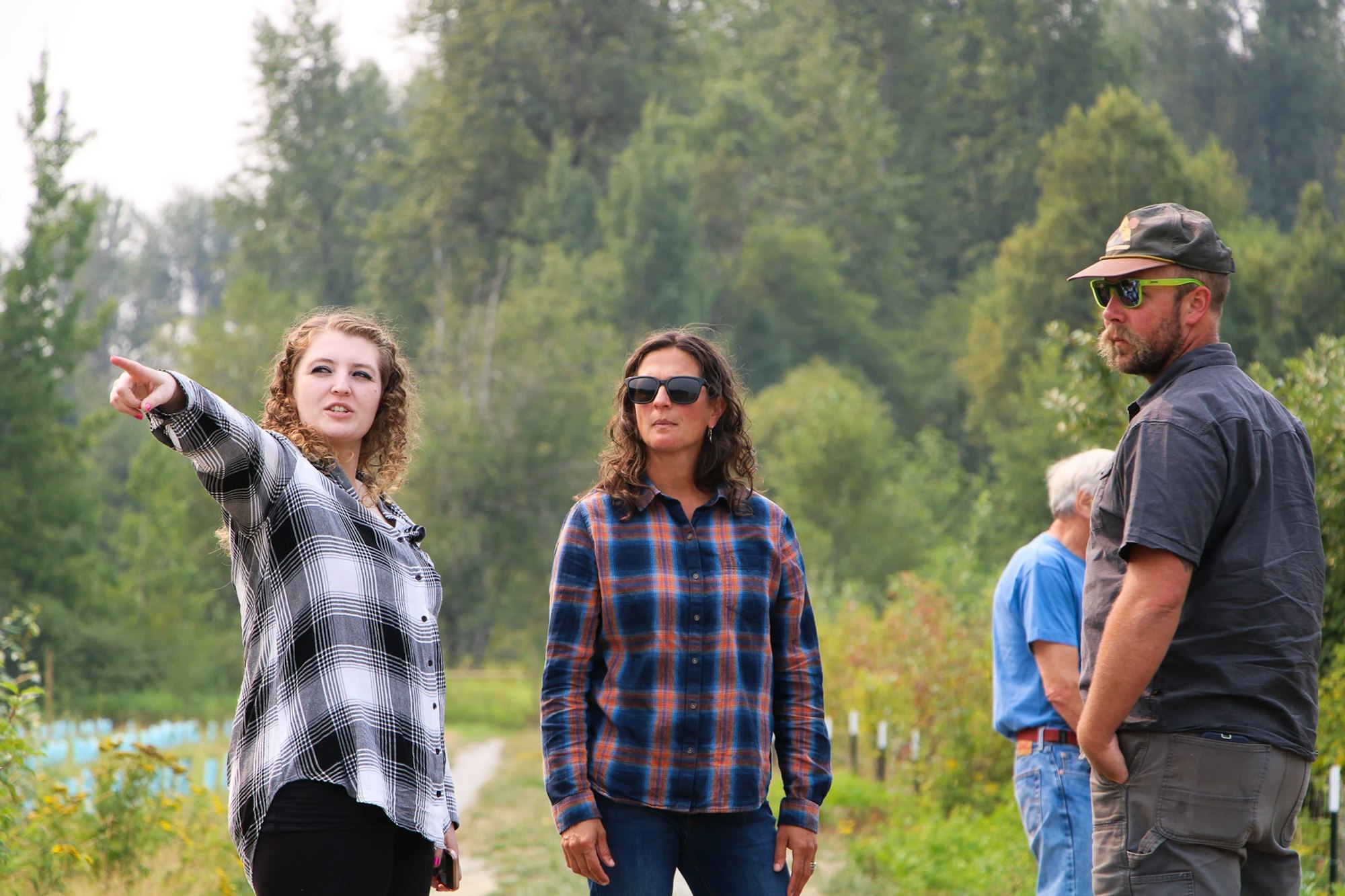 Three people stand outdoors; a woman in plaid points ahead, another in sunglasses observes, and a man with a cap watches.
