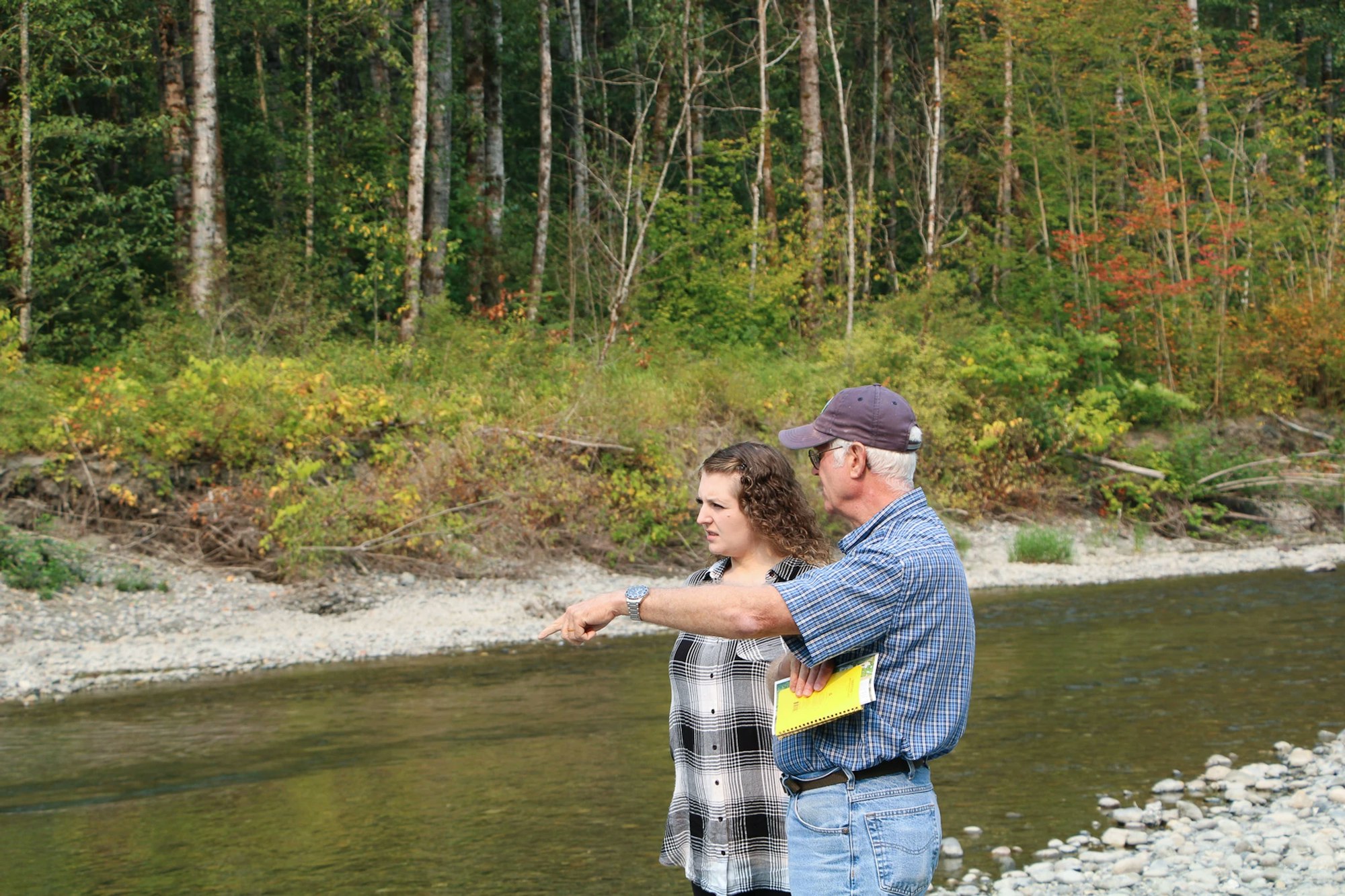 Two people standing by a riverbank in a forested area, one pointing outward.
