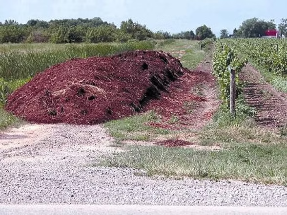 A large pile of red mulch beside a gravel path, surrounded by greenery and fields.