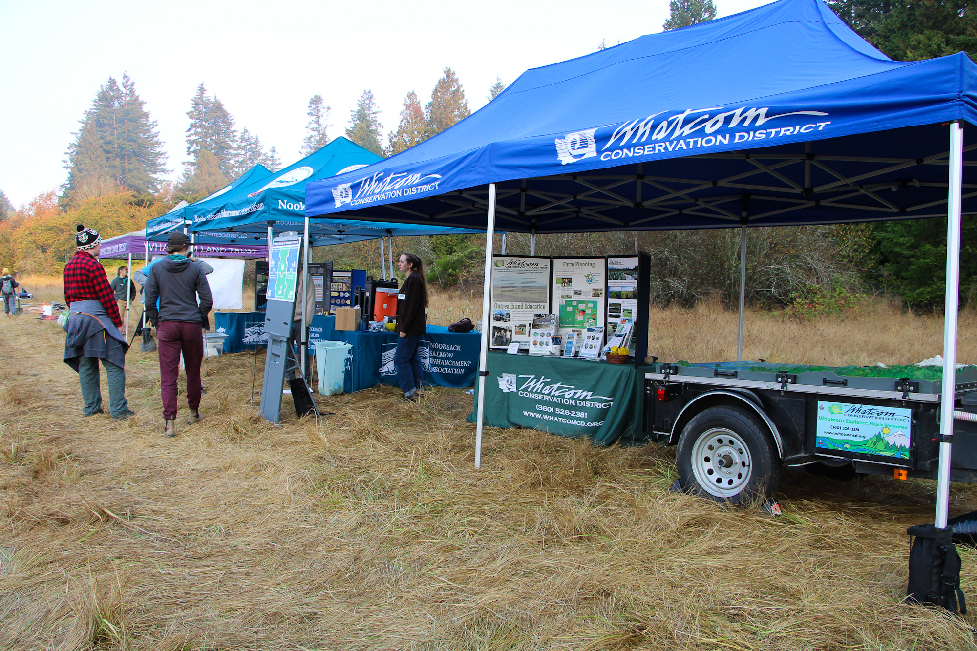 Outdoor event with blue tents labeled "Whatcom Conservation District," field displays, and people attending.